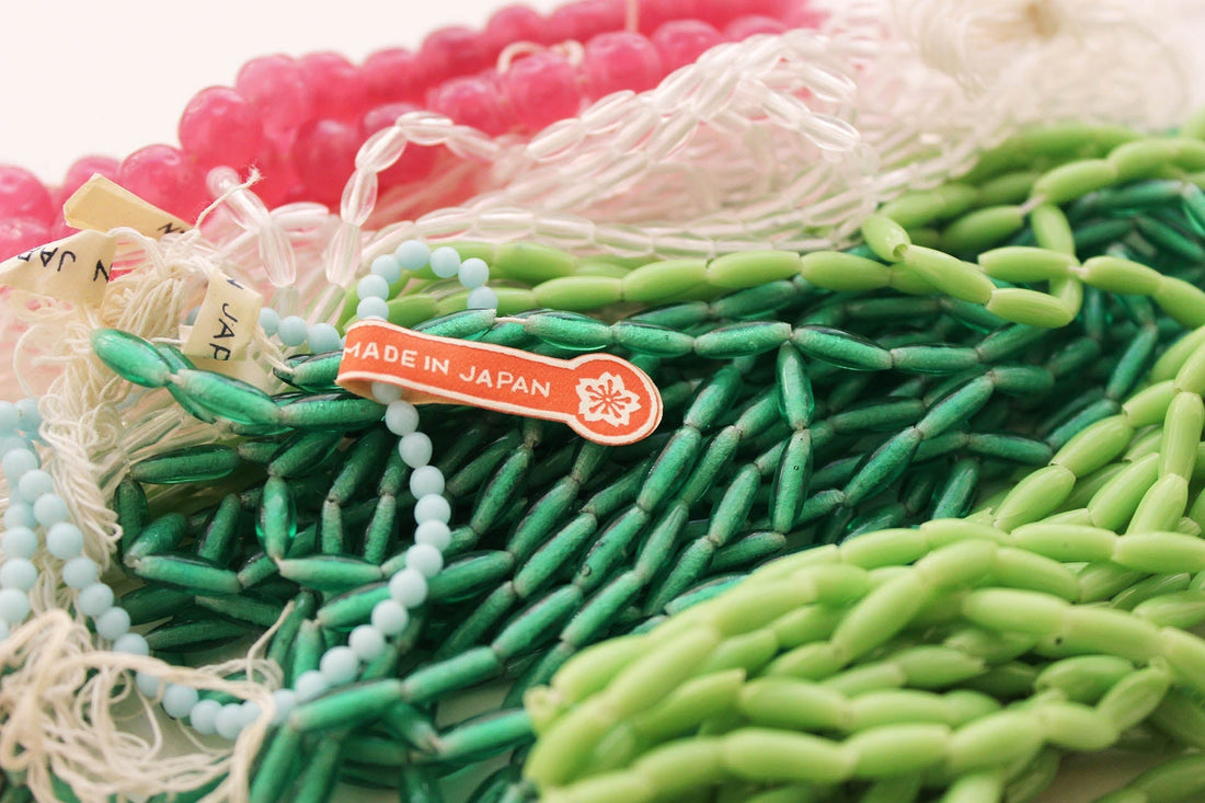 Strands of vintage Japanese beads in shades of pink, white, mint and emerald green, with both round and elongated shapes, are gathered together. A red “Made in Japan” tag and small handwritten labels highlight their origin.
