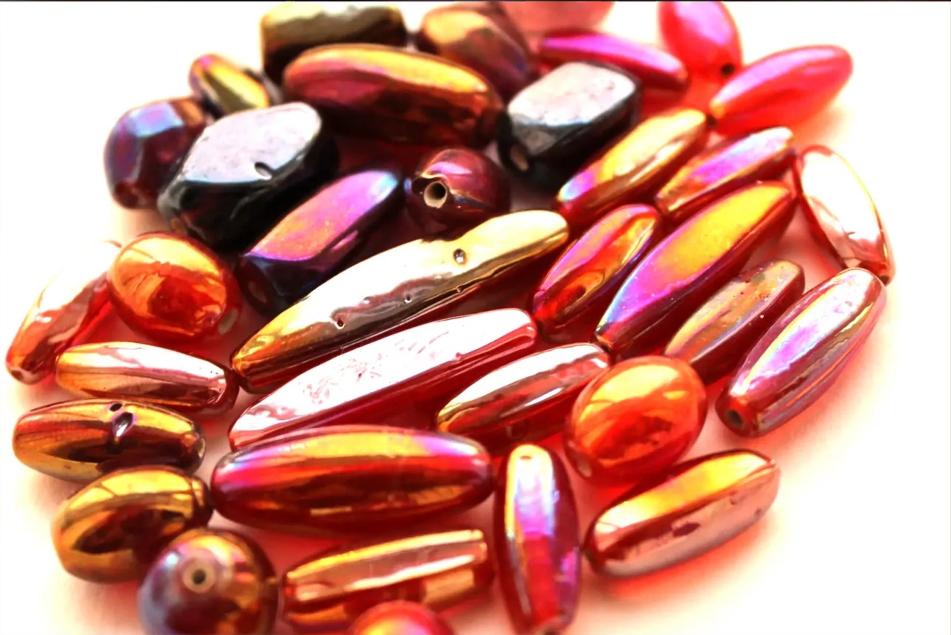 A close-up of numerous iridescent glass beads in shades of deep red, orange, gold, and magenta. The beads vary in shape — some are long and slender, others are oval or irregular — and they are scattered in a loose cluster on a light, neutral surface.