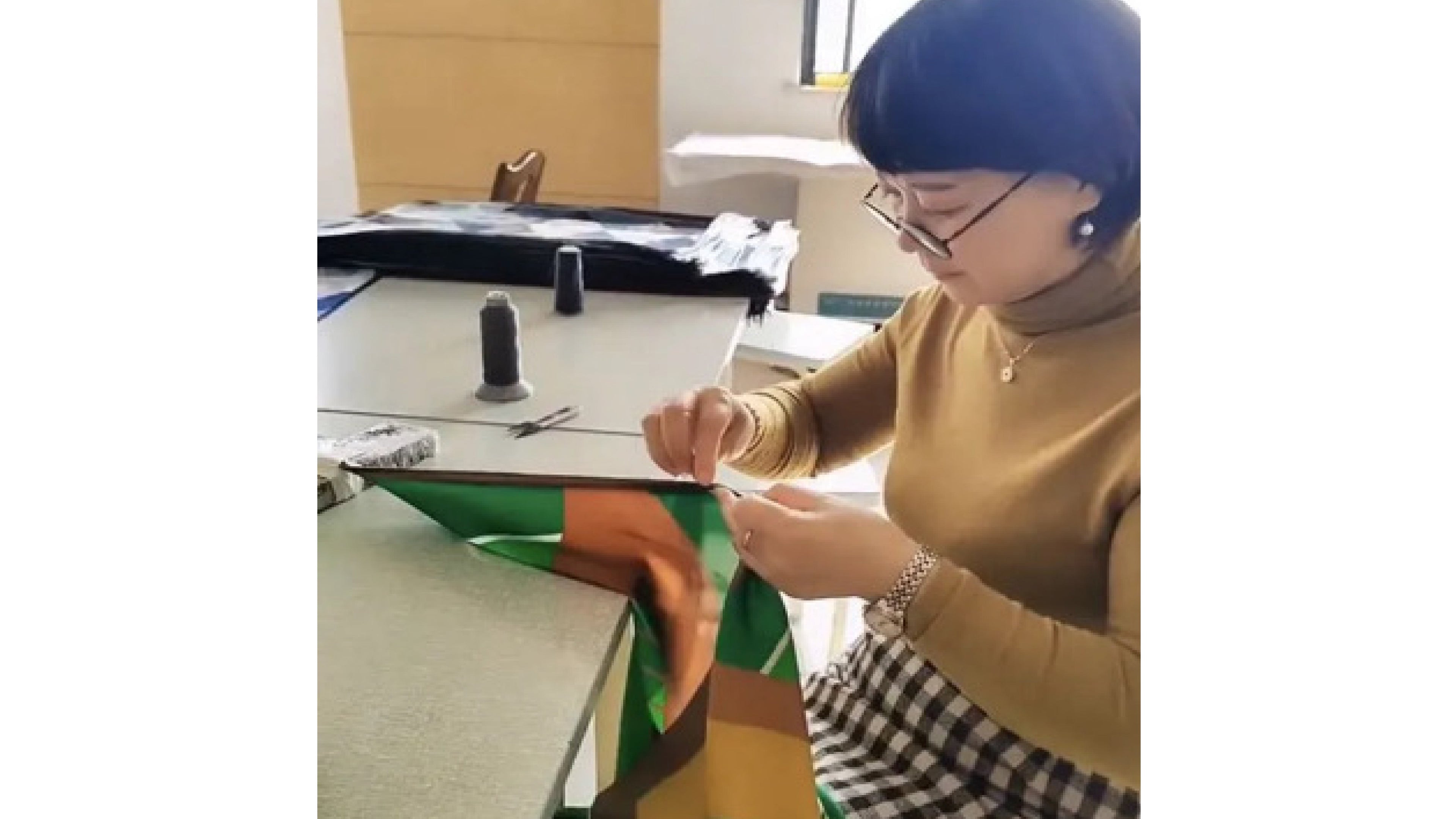 A woman in glasses and a turtleneck hand-sews colorful fabric at a sewing table.